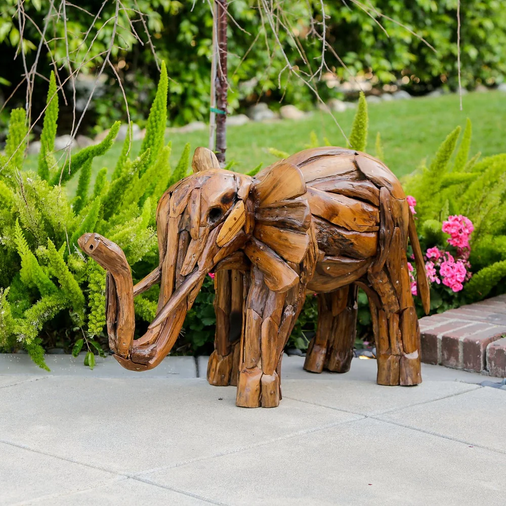 A carved wooden elephant sculpture in a garden, surrounded by green plants and pink flowers.