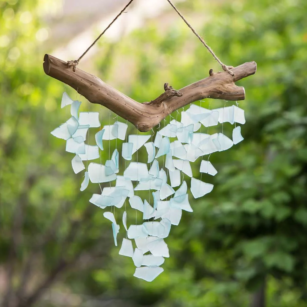 A rustic wooden hanging wind chime adorned with white and light blue glass pieces, suspended outdoors against a lush green background.